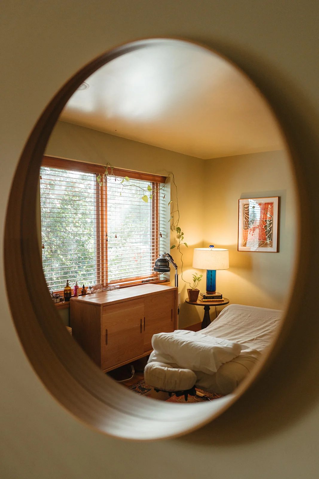 A round wooden mirror reflects a bright treatment room with windows, a lamp and a made table.