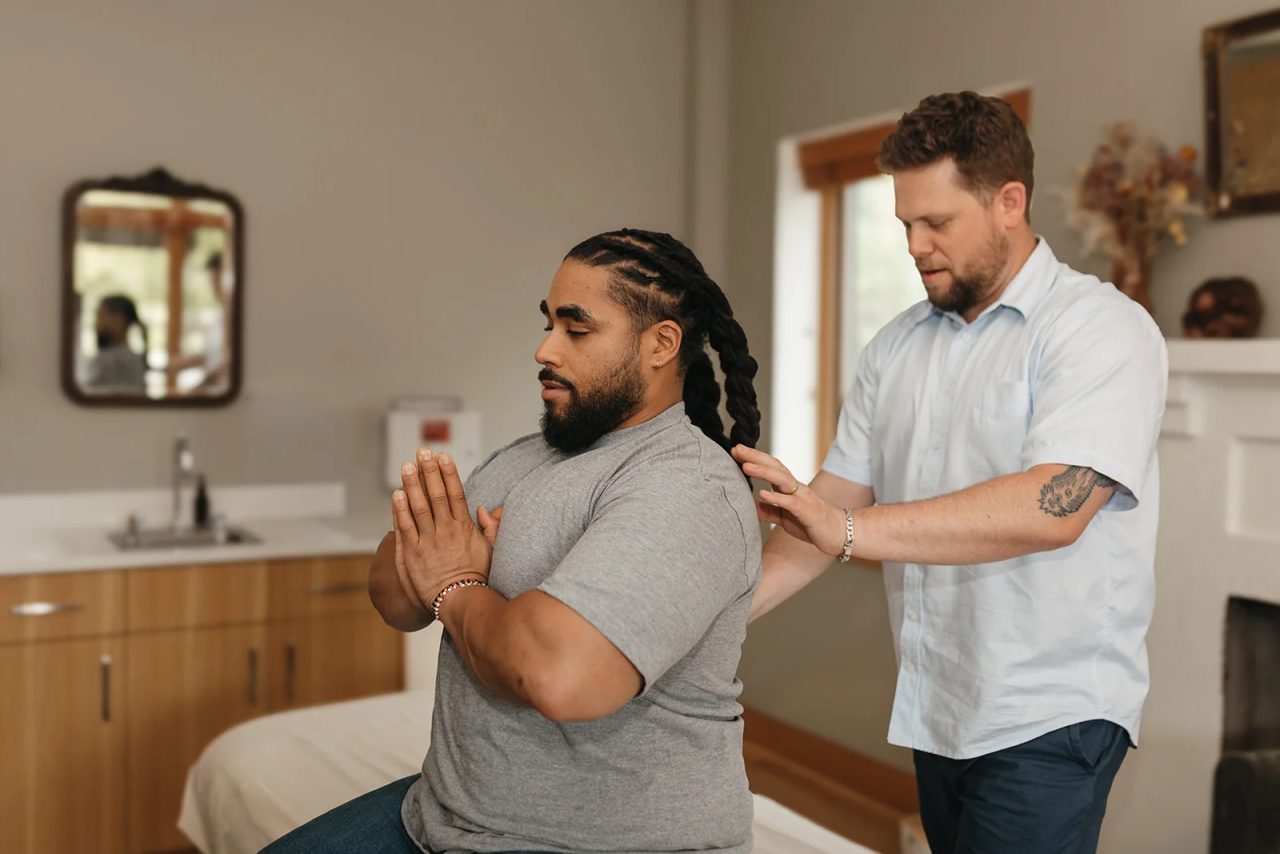 A practitioner rests his hands on a seated patient's shoulders while the patient holds his palms together.