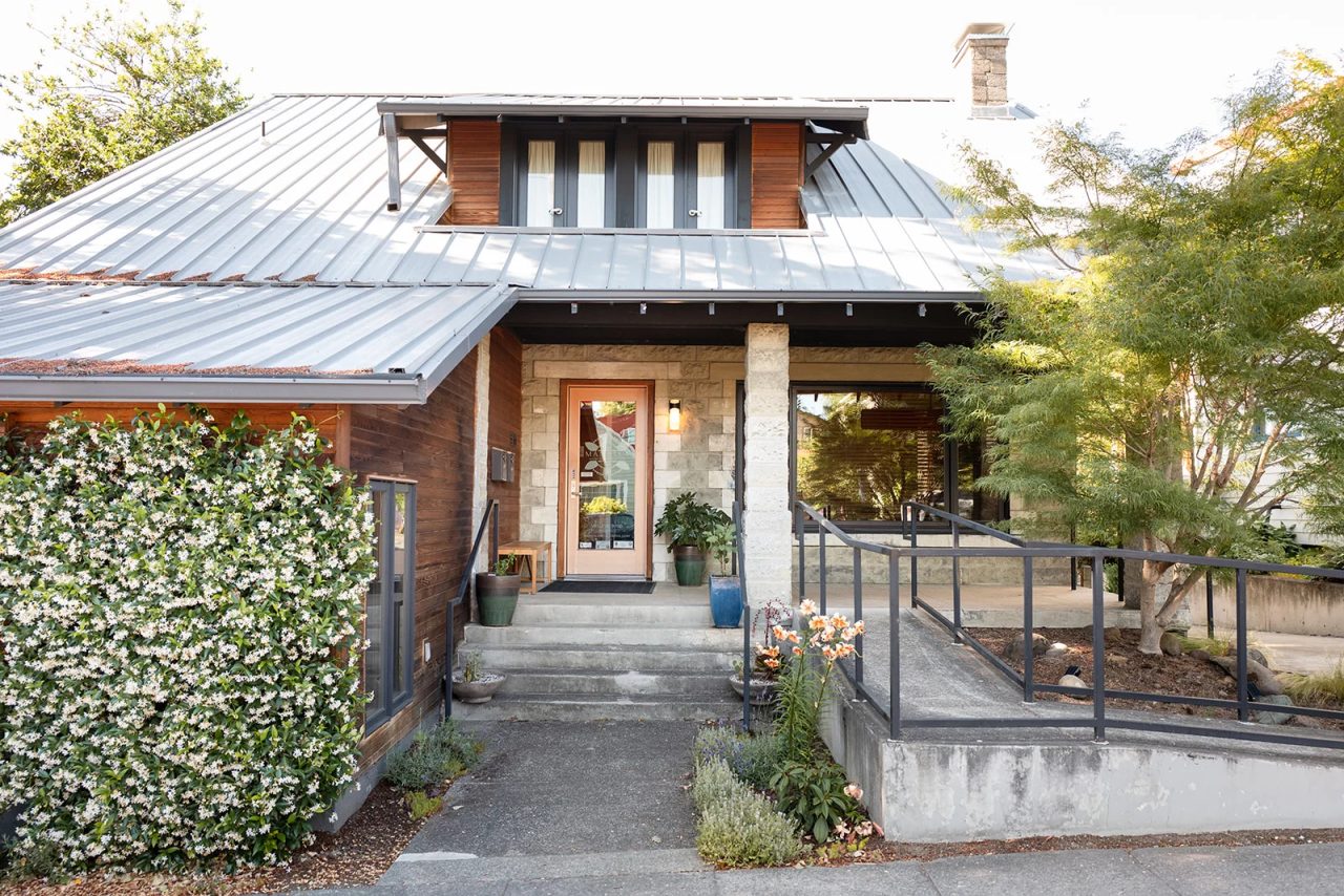 A wider view of the clinic's front: the metal roof and dormer, the jasmine wall, the steps, the ramp and the stone porch.