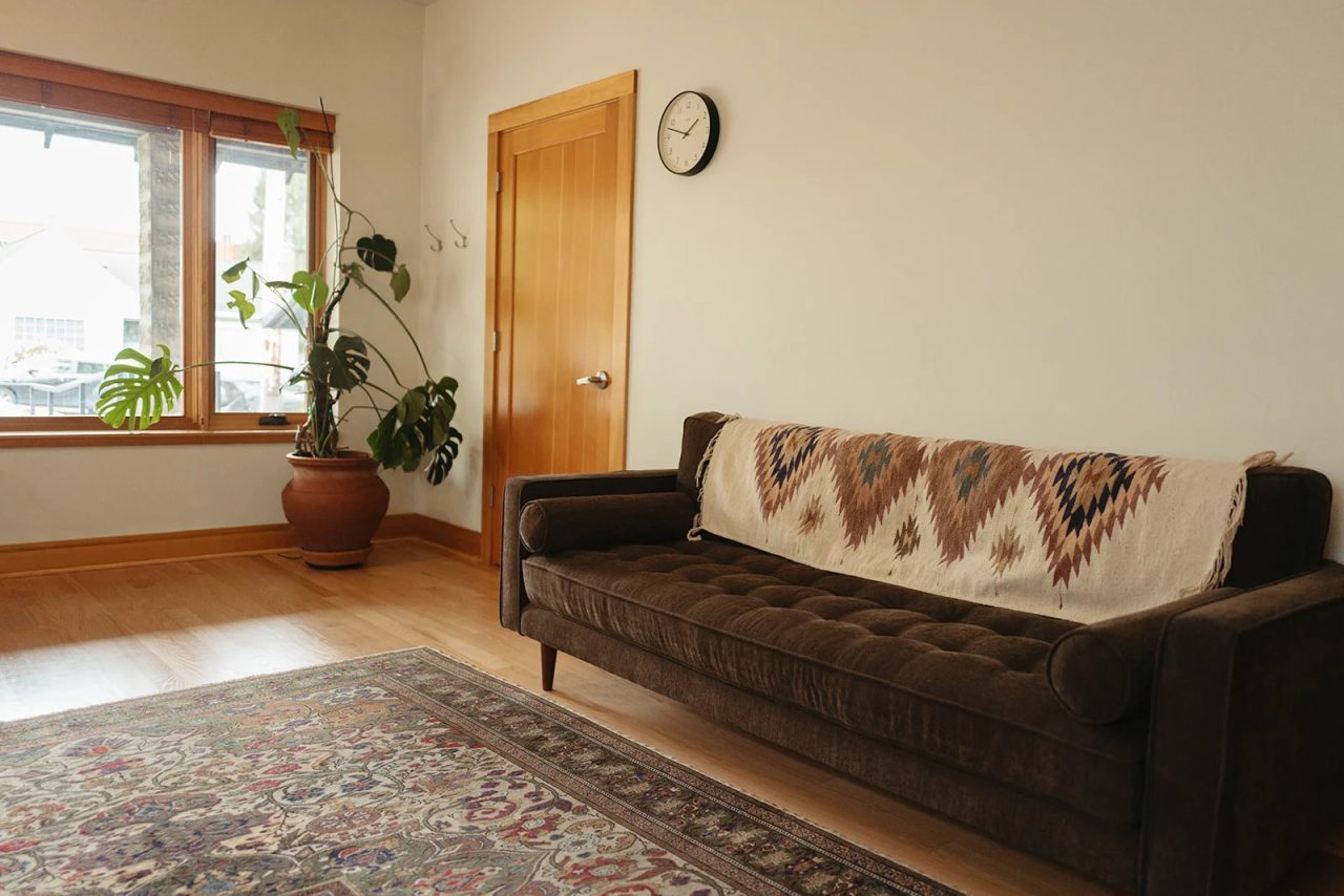 A counseling room with a brown tufted sofa under a patterned throw, a patterned rug, a monstera by the window and a wall clock.
