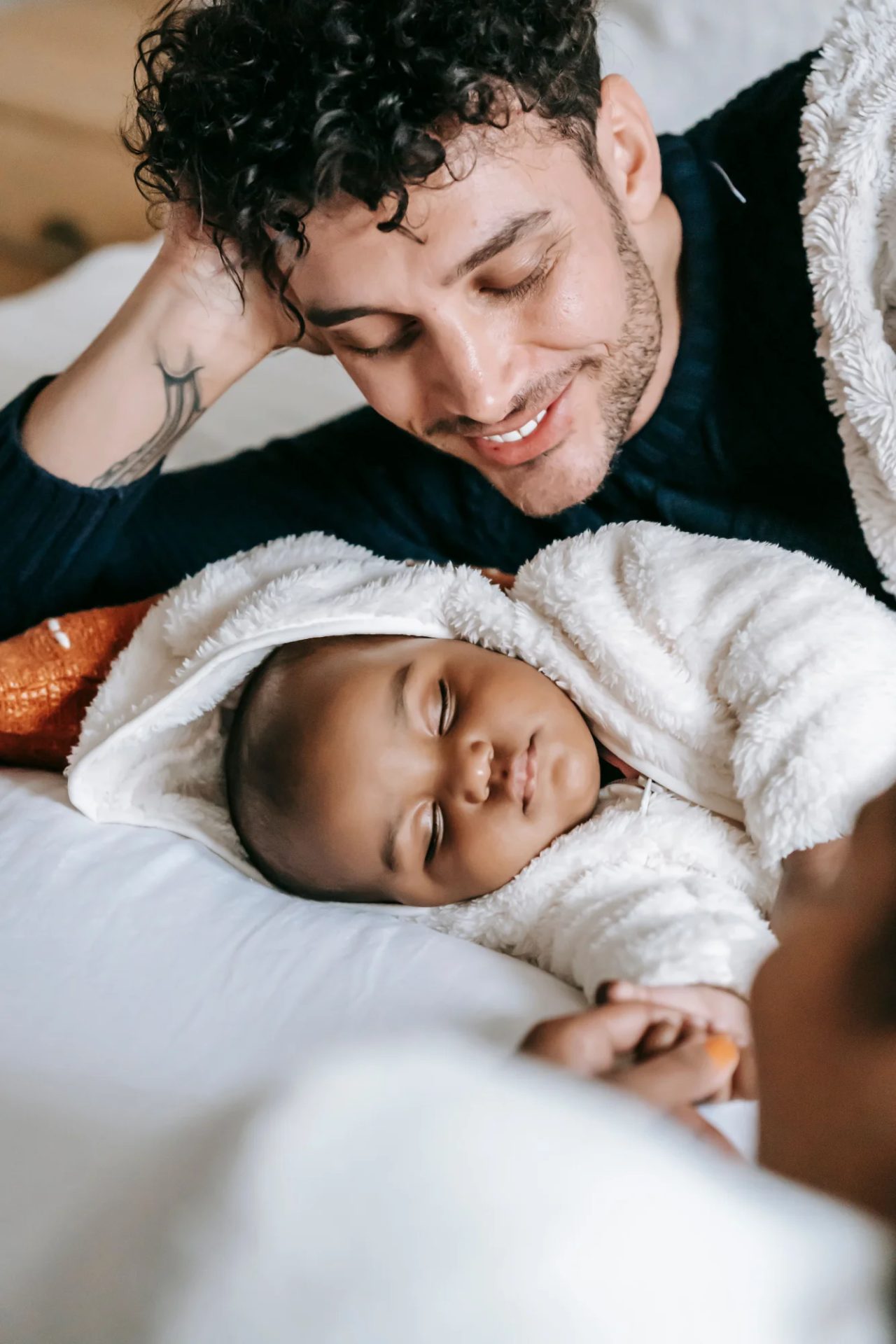 A father lies on a bed looking down at his sleeping newborn, wrapped in a white blanket.