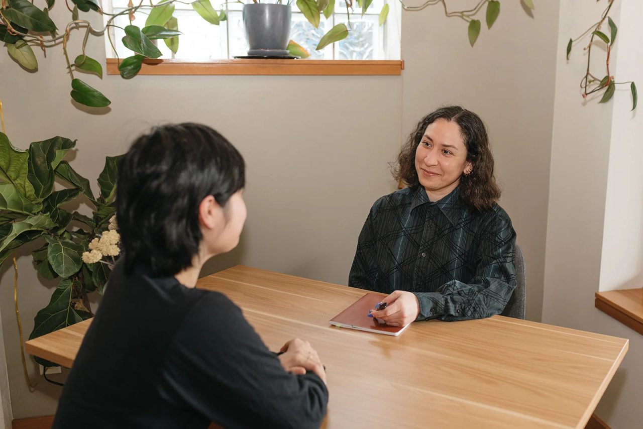 A practitioner listens to a patient across a wooden table in a bright room with trailing plants.