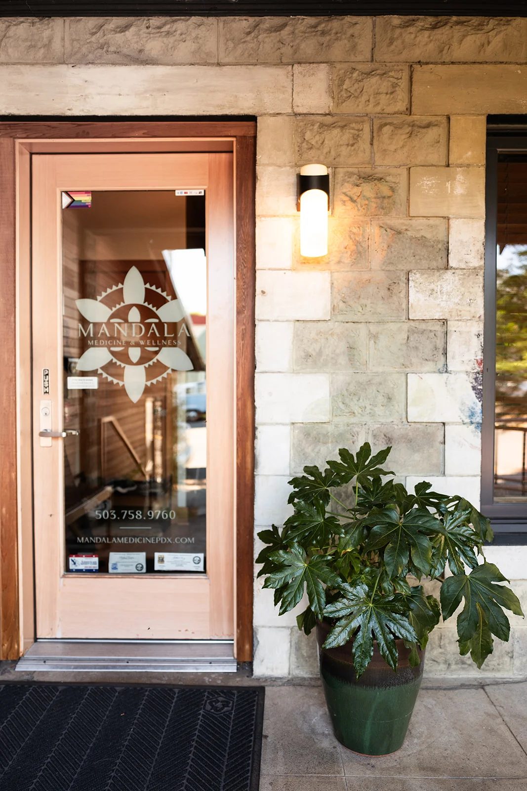 The clinic's glass front door with the Mandala logo, beside a stone wall, a wall lamp and a potted plant.