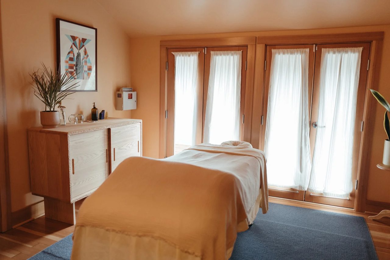 A large treatment room with a made table on a blue rug, a wooden dresser, a framed print and sheer curtains over French doors.