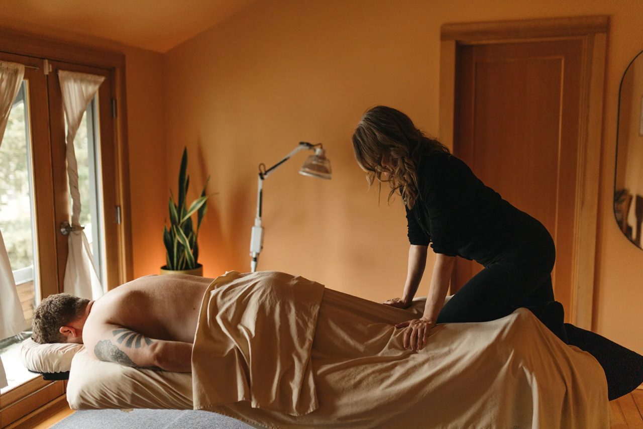 A massage therapist works along a man's back as he lies face down, in a warm room with French doors and a floor lamp.