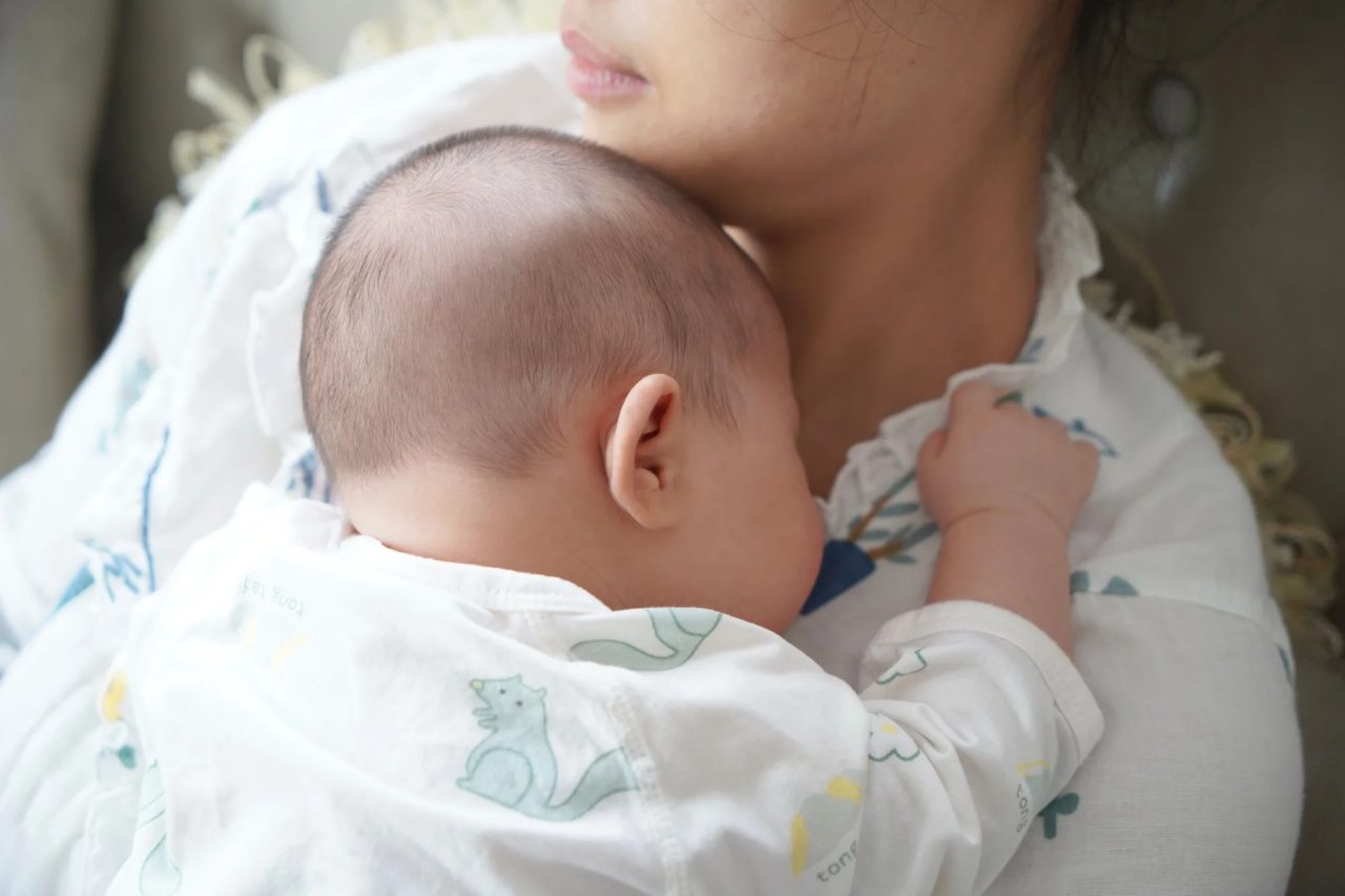 A newborn in a patterned swaddle rests skin-to-skin against a mother's shoulder.