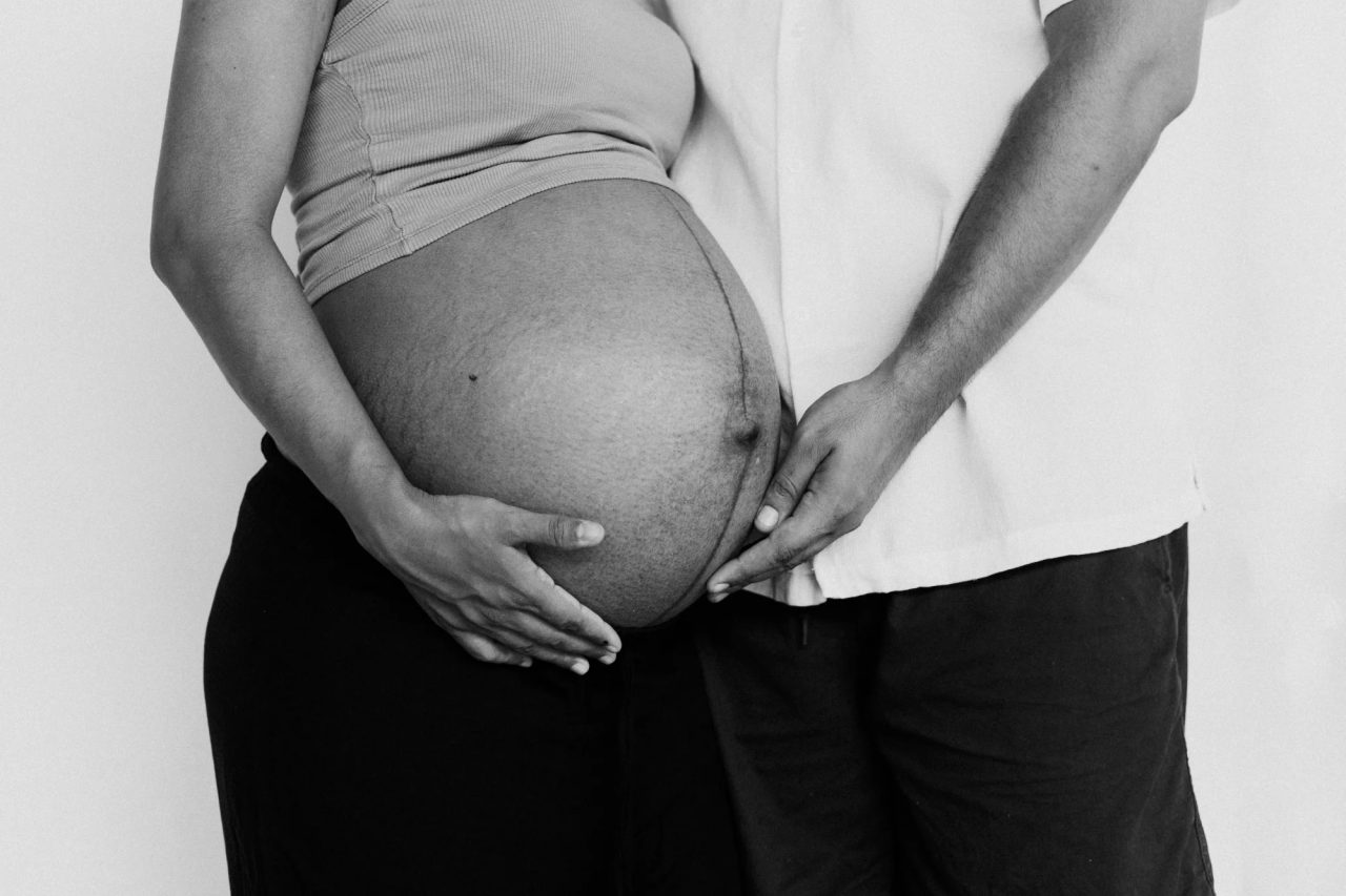 Black-and-white close-up of a pregnant woman's hands and her partner's hand resting together on her belly.