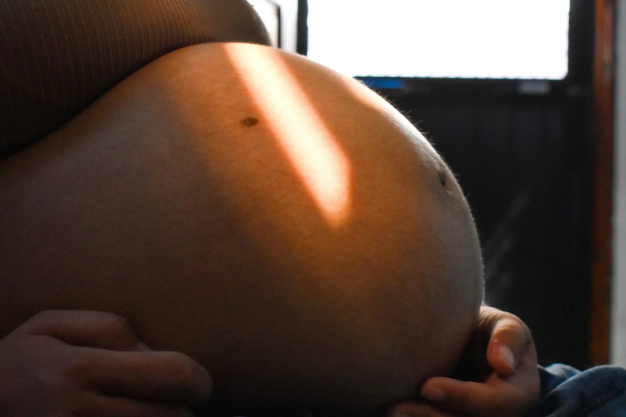 A pregnant belly in a stripe of warm window light, one hand resting on it.