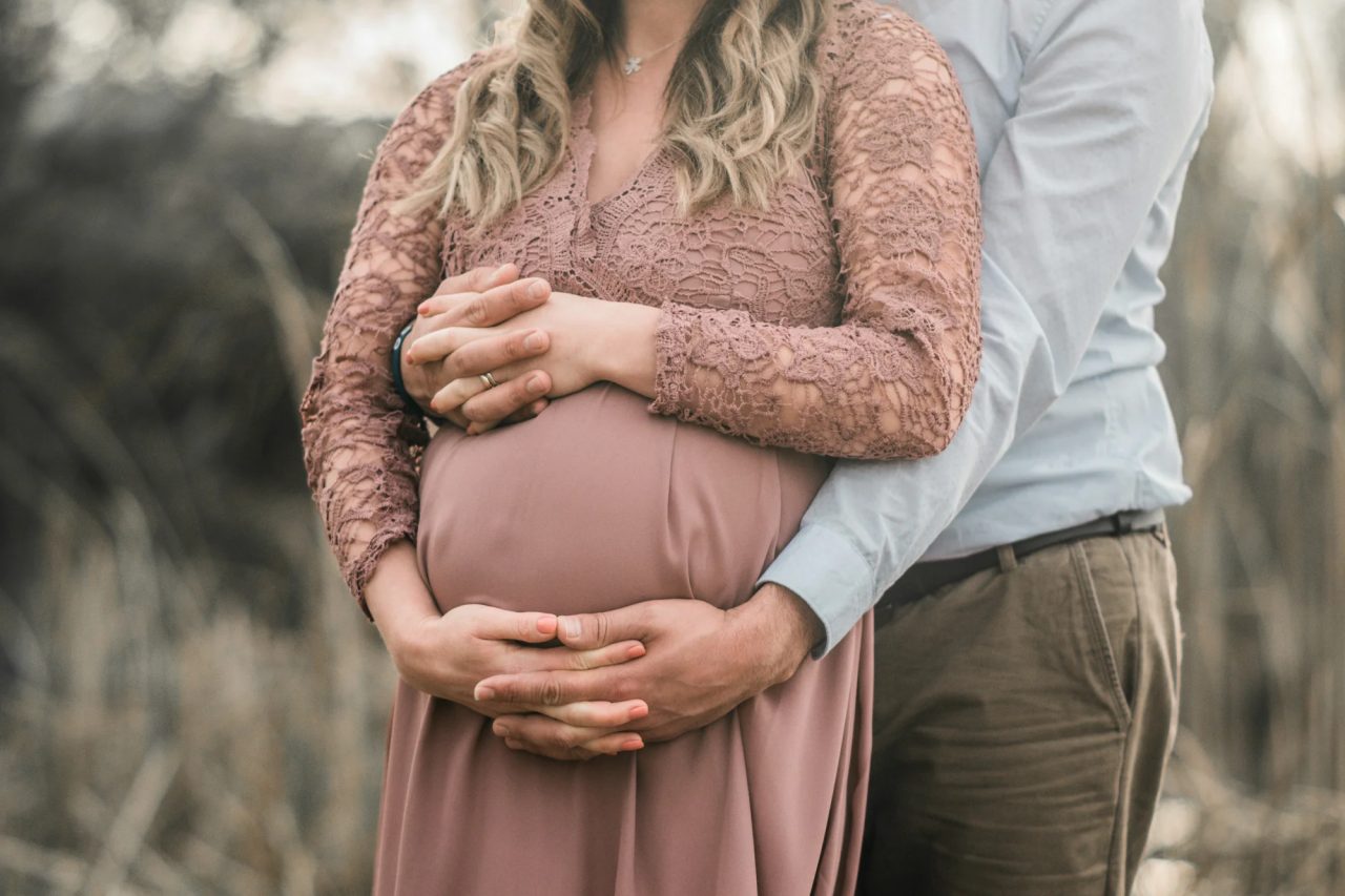 A pregnant woman in a lace dress stands in tall backlit grass, her partner's hands clasped with hers over her belly.