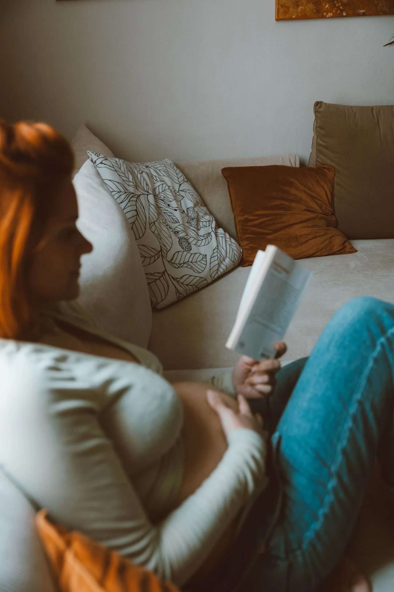 A pregnant woman reclines on a couch with a book in a sunlit living room.