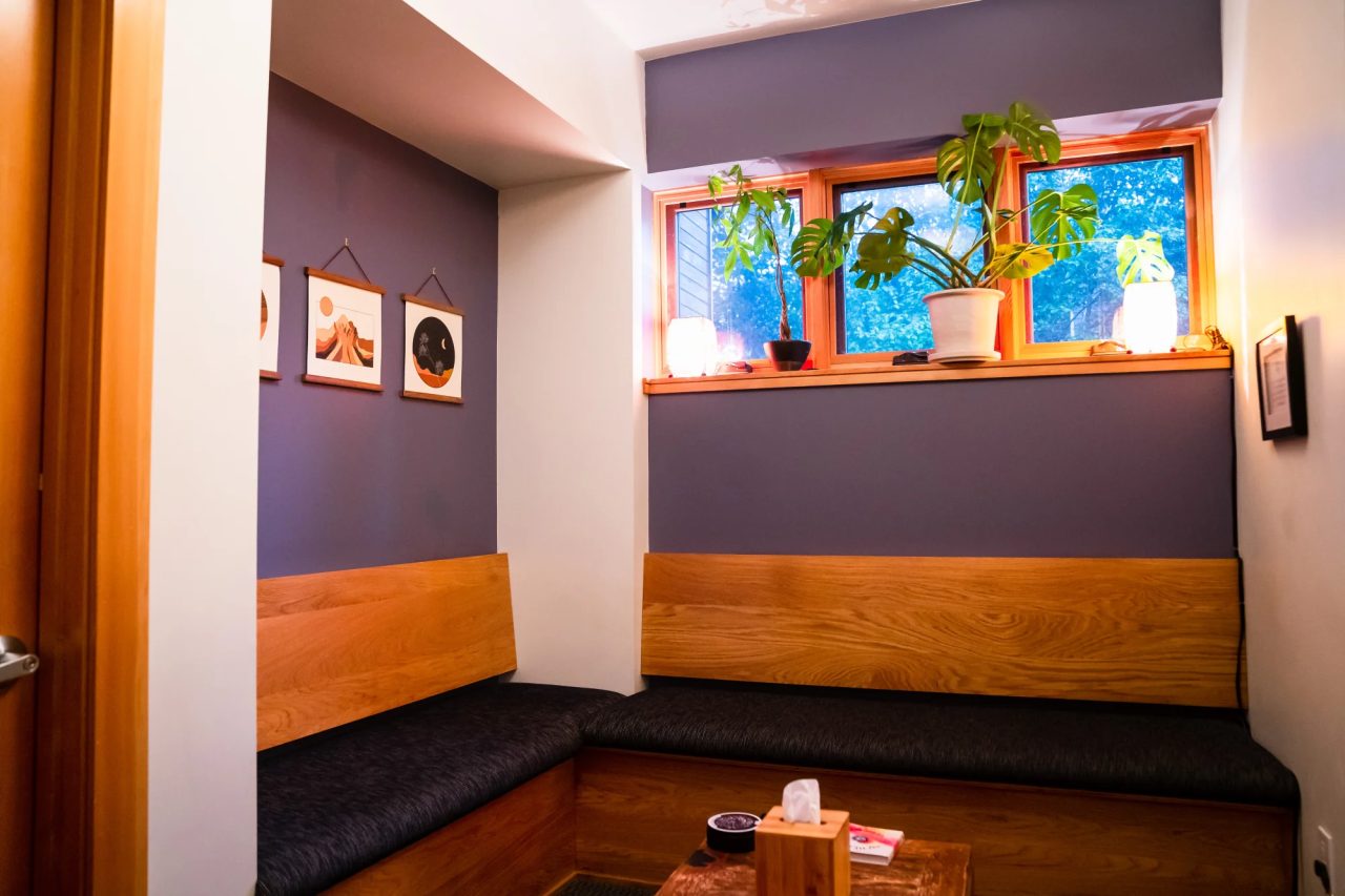 A waiting nook with cushioned wooden benches along two walls, three framed prints, and plants on a sunny windowsill.