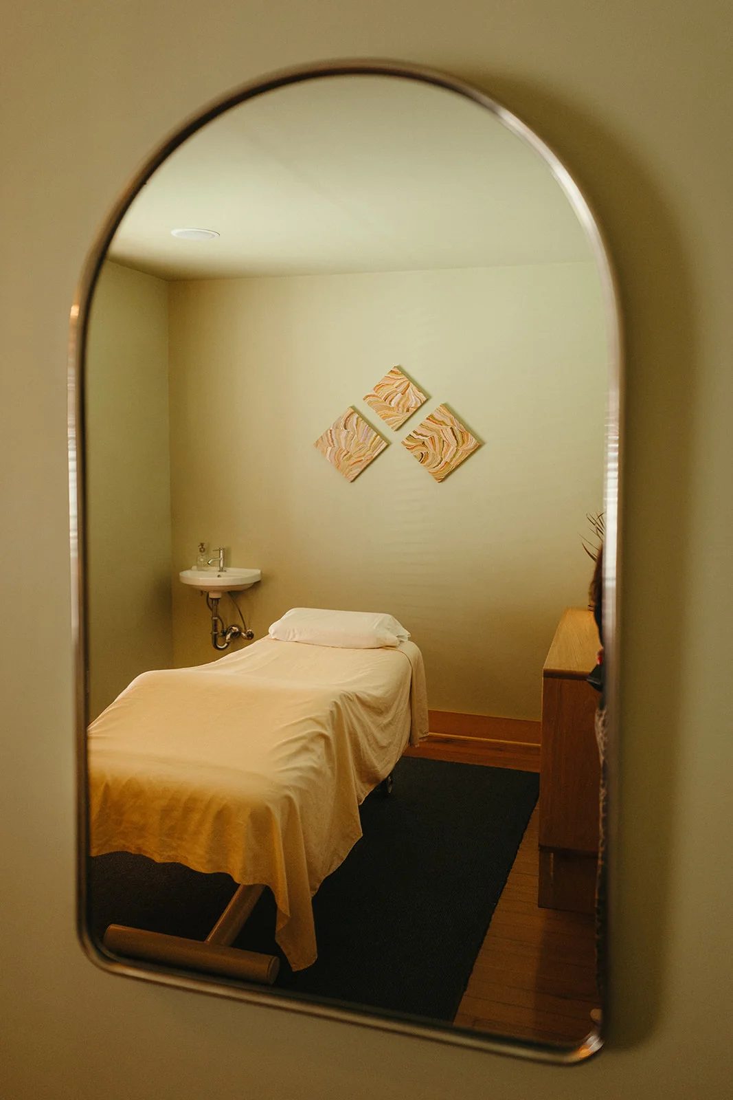 An arched mirror reflects a treatment room with a made table, a small sink and wooden tiles on the wall.