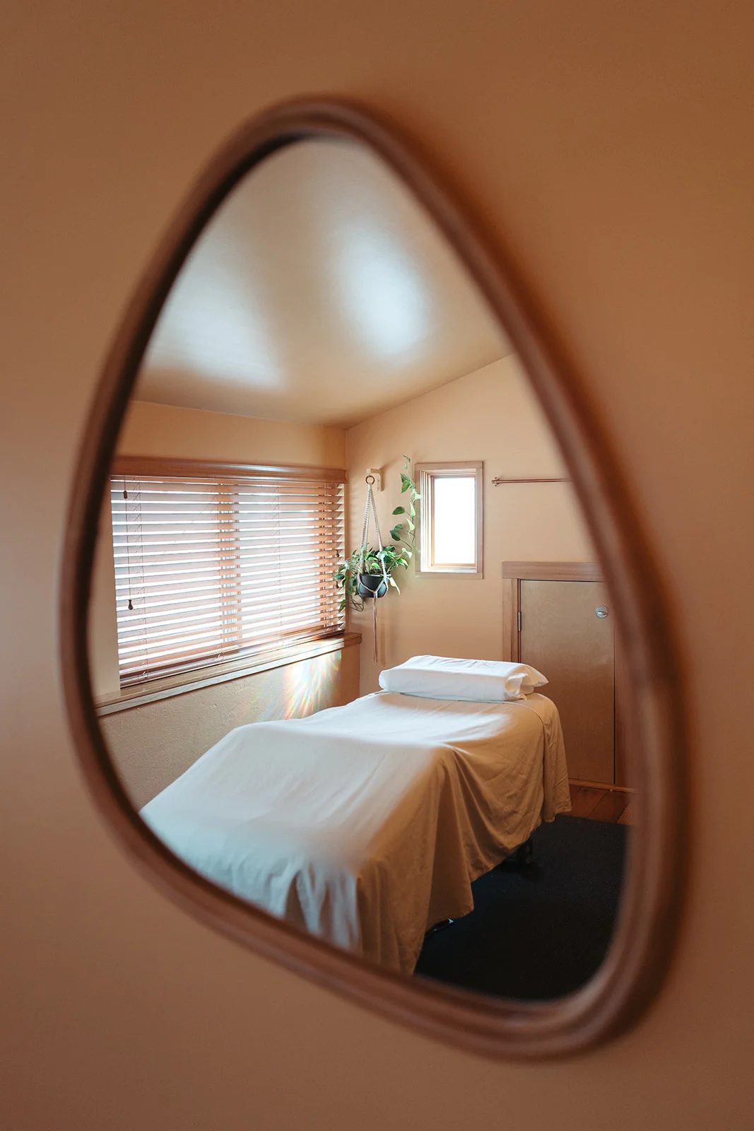 A treatment room reflected in a curved wooden mirror: a made table under a window with blinds.