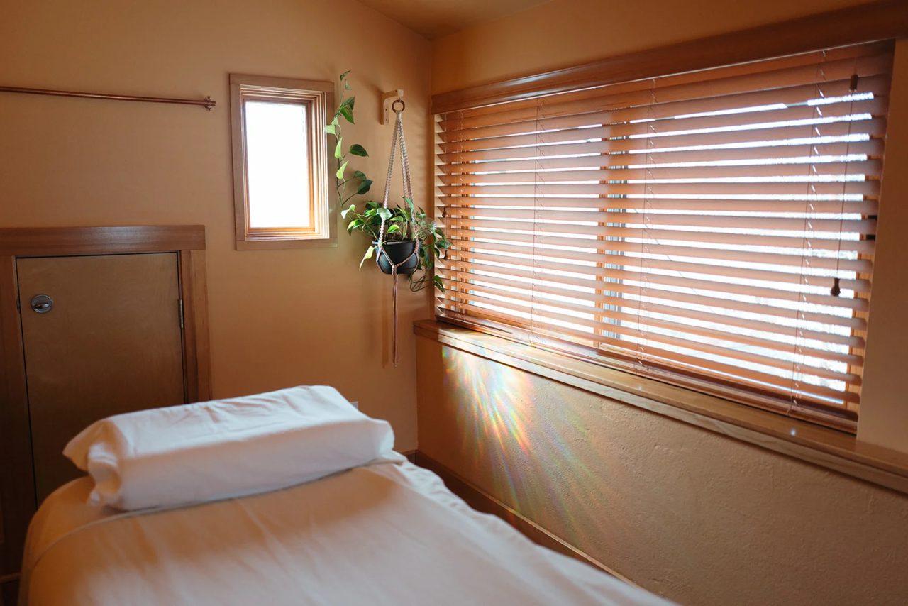A quiet treatment room: a made table under a wide window with wooden blinds, a hanging plant, and small rainbows of light on the wall.