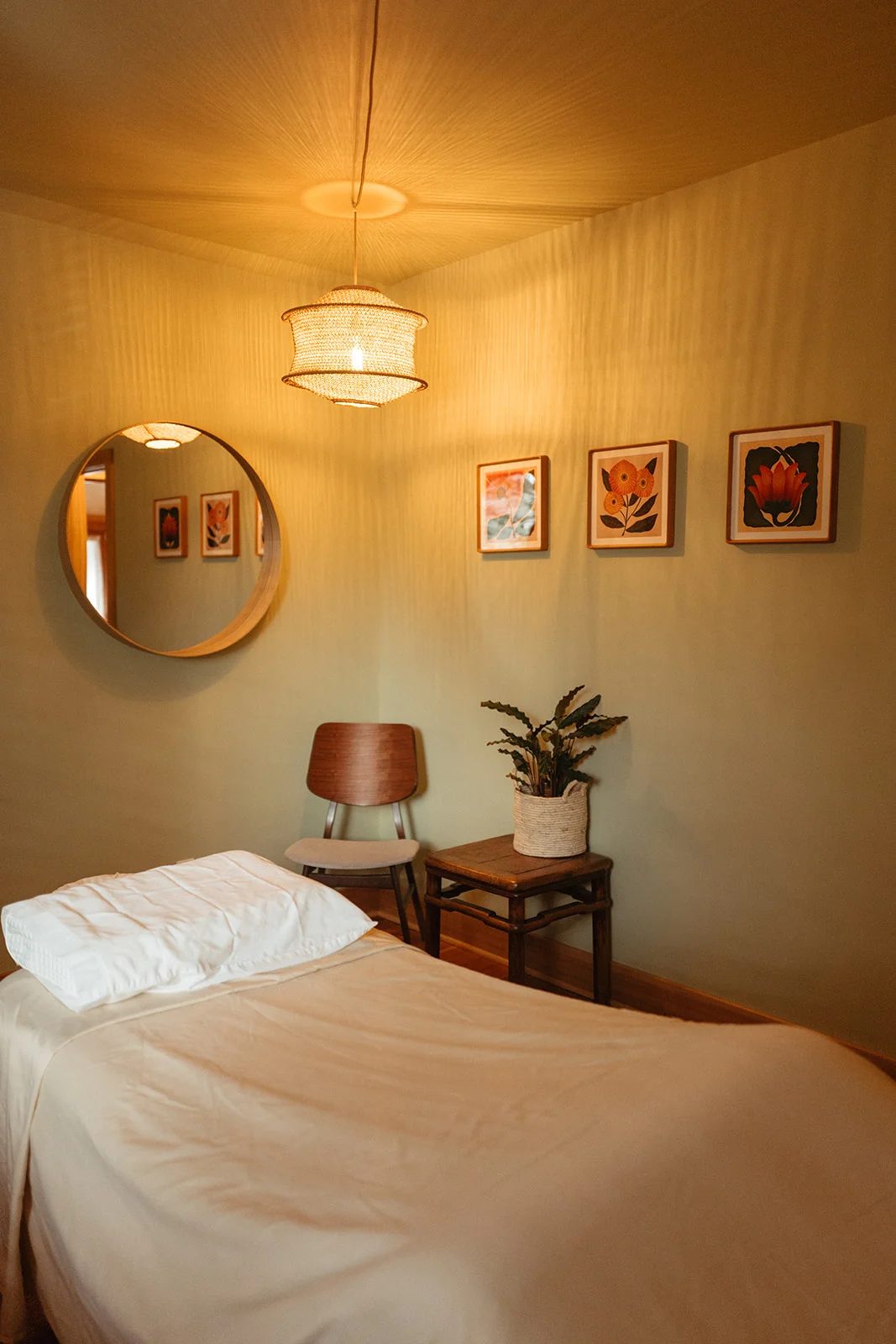 A treatment room with a woven pendant lamp, a round mirror, three framed flower prints, a chair and a made table.